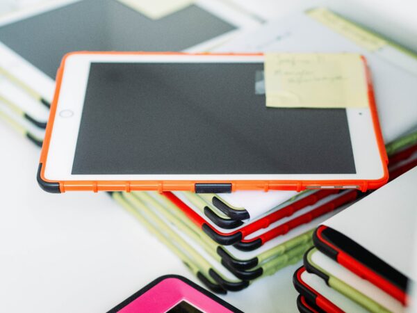 I pad with orange case, ontop of a stack of multiple other I pads with varying case colours, which are green and pink.